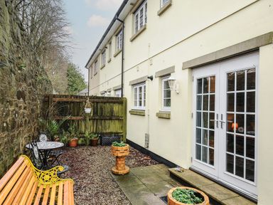 A courtyard with a yellow bench a small black metal table with two chairs potted plants and white double doors at 1 Bethany Court in Bodmin