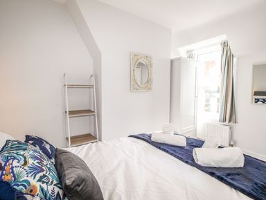 A bedroom with a bed, pillows, rolled towels, a ladder shelf, a white wardrobe, a window with curtains, and a mirror on the wall at Quarrier's Rest in Swanage