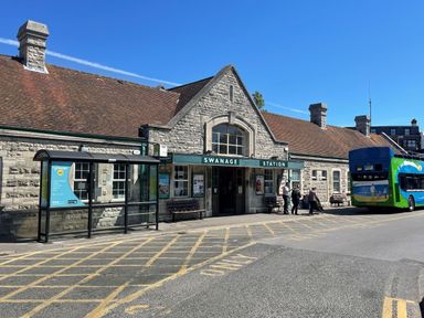 The exterior of Swanage Station with a bus stop and people near a green and blue bus at Quarrier's Rest in Swanage