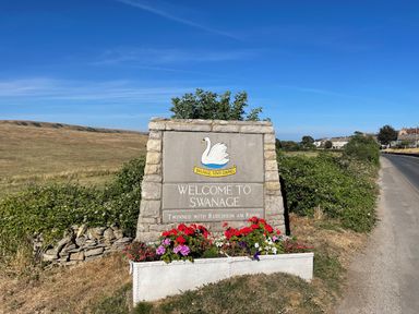 A stone welcome sign with flowers next to a road and grass fields at Quarrier's Rest in Swanage