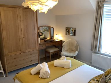 A bedroom with a wooden wardrobe a dressing table with mirror and lamp a white chair with fur and window with striped curtains at Quarrier's Rest in Swanage