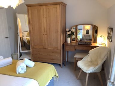 A bedroom with a bed with rolled towels on a yellow blanket a wooden wardrobe a dressing table with mirror and lamp and a white chair with a fur throw at Quarrier's Rest in Swanage