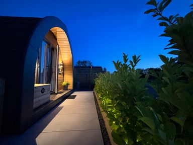 An arched wooden cabin entrance with a lit potted plant by a pathway and green bushes with lights at Oh Deer in Ashbourne