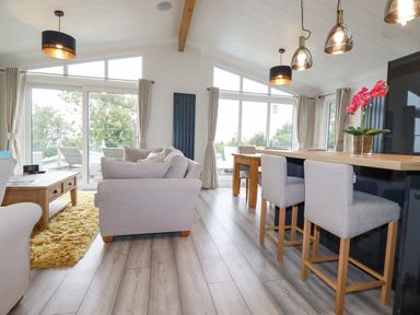 A living room and kitchen area with light gray sofas wooden tables and chairs and large windows at Mordros Lodge in St Day