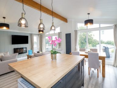 An open plan living and dining area with a wooden island table and pink flowers a dining table with chairs large windows and a mounted television at Mordros Lodge in St Day