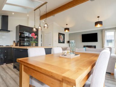 A dining table with chairs near a kitchen island with flowers and a living room with a wall-mounted TV at Mordros Lodge in St Day