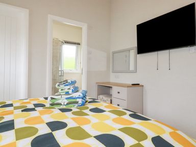 A bedroom with a bed covered in a colorful patterned bedspread with folded towels on top a dresser with a mirror and a wall mounted television at Mordros Lodge in St Day
