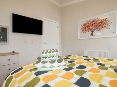 A bedroom with a bed with patterned bedding and towels on it a wall-mounted tv a chest of drawers a door and a framed artwork at Mordros Lodge in St Day