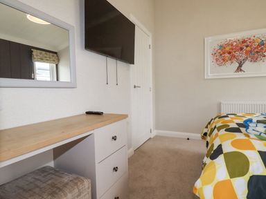 A bedroom with a bed featuring a geometric patterned duvet a wall-mounted TV a dresser with a remote a mirror and a framed picture of a tree at Mordros Lodge in St Day