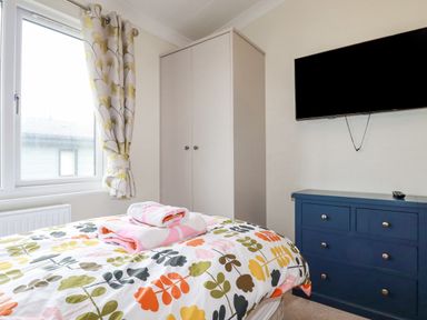 A bedroom with a bed with colorful leaf patterned bedding a window with curtains a white wardrobe a blue chest of drawers and a wall mounted TV at Mordros Lodge in St Day
