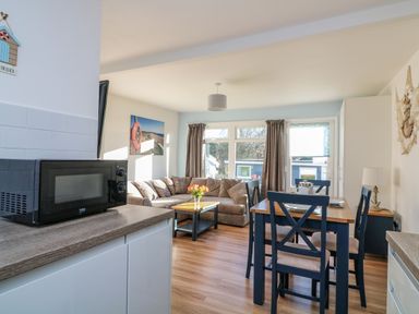 A dining area with a wooden table and four chairs next to a living room with a corner sofa and windows at Beachcomber in Norton Park near Dartmouth