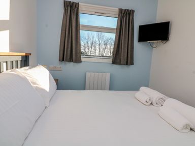 A bedroom with a bed covered in white linens towels on the bed a window with curtains and a wall-mounted television at Beachcomber in Norton Park near Dartmouth