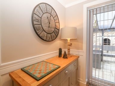 A wooden sideboard with a checkered board lamp and two decorative stones under a wall clock next to a window with vertical blinds at Ty Bains in Llanberis