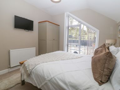 A bedroom with a bed white bedding and brown pillows a wardrobe a wall mounted television and a window with vertical blinds at Ty Bains in Llanberis