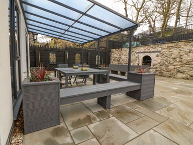 An outdoor patio area with a glass roof, a gray dining table with chairs, built-in benches with planters, and a stone wall with a built-in fireplace at Ty Bains in Llanberis