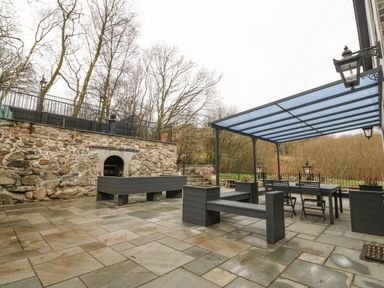 An outdoor patio with stone flooring a covered seating area a stone wall with fireplace and trees in the background at Ty Bains in Llanberis