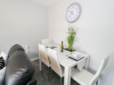 A dining area with a white table and four white chairs a bottle of wine and plates on the table a black leather couch and a wall clock at 17 Ffordd Gwenllian in Llanfairpwllgwyngyll