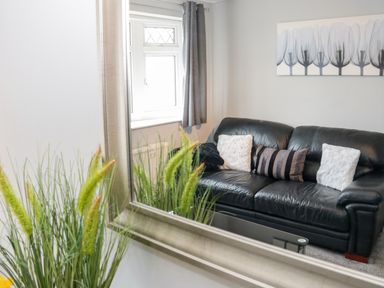 A black leather sofa with pillows in front of a window with gray curtains and a floral painting on the wall at 17 Ffordd Gwenllian in Llanfairpwllgwyngyll