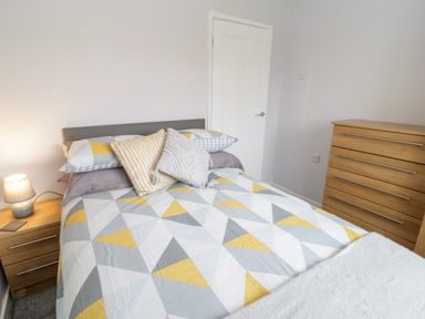 A bedroom with a bed covered in geometric patterned bedding two cushions two wooden dressers and a lamp on one dresser at 17 Ffordd Gwenllian in Llanfairpwllgwyngyll