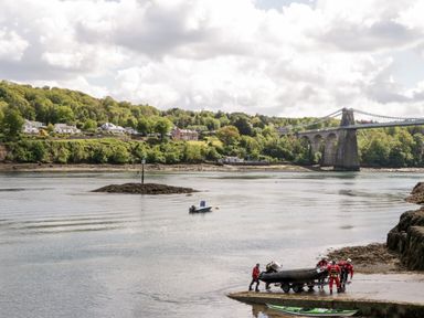 A river scene with a group of people loading a boat on a trailer near a stone embankment and a suspension bridge in Llanfairpwllgwyngyll