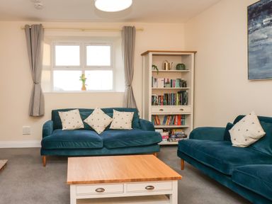 A living room with two blue sofas a wooden coffee table a bookshelf and a window with curtains at Garden Cottage in Alnmouth