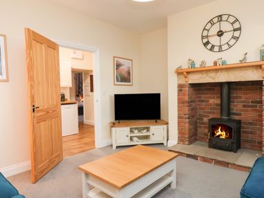 A living room with a wooden coffee table a TV on a stand an open wooden door and a brick fireplace with a fire burning at Garden Cottage in Alnmouth