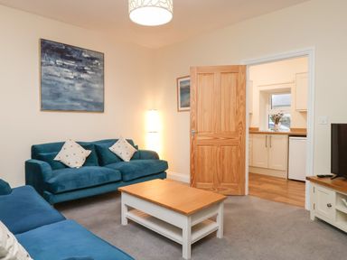 A living room with blue sofas a wooden coffee table and an open door leading to a kitchen at Garden Cottage in Alnmouth
