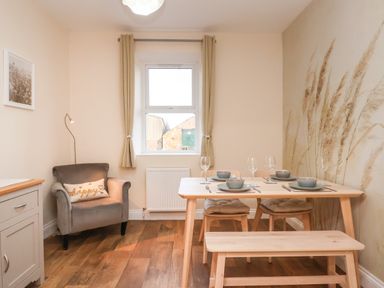 A dining room with a wooden dining table set with dishes and glasses a gray armchair with a pillow and a window with curtains at Garden Cottage in Alnmouth