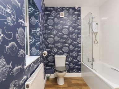 A bathroom with a toilet and bathtub with shower and fish patterned wallpaper at Garden Cottage in Alnmouth