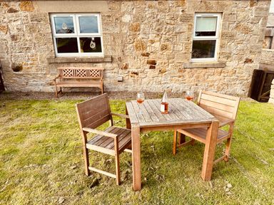A wooden table with two chairs and wine glasses on grass outside a stone house at Garden Cottage in Alnmouth