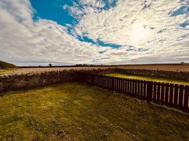 A grass yard enclosed by a stone wall and wooden fence under a partly cloudy sky at Garden Cottage in Alnmouth