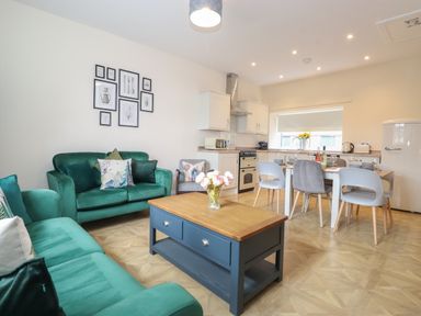 A combined living room and kitchen with green sofas a wooden coffee table dining table with chairs and kitchen appliances at Pond Cottage in Alnmouth