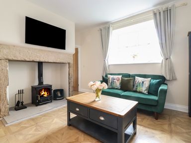 A living room with a green sofa, coffee table with flowers, wood burning stove, and wall-mounted television at Pond Cottage in Alnmouth