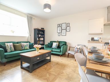 A living room with green sofas a coffee table a bookshelf wall art and a dining table at Pond Cottage in Alnmouth