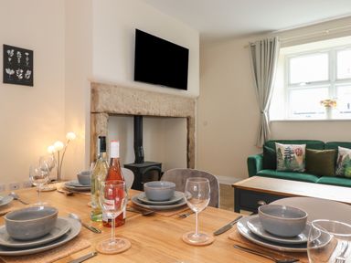 A dining table set with plates bowls wine glasses and bottles in a living room with a stone fireplace a wood stove a green sofa and window at Pond Cottage in Alnmouth