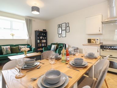 A dining table set with grey bowls plates and wine glasses in a room with green sofas a bookshelf and a kitchen area at Pond Cottage in Alnmouth