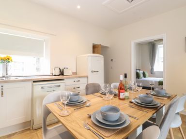 A kitchen dining area with a wooden table set with plates bowls cutlery and wine glasses beside a fridge and window at Pond Cottage in Alnmouth
