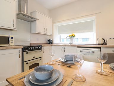 A kitchen with white cabinets a stove a microwave toaster kettle and a dining table set with plates bowls glasses and cutlery at Pond Cottage in Alnmouth