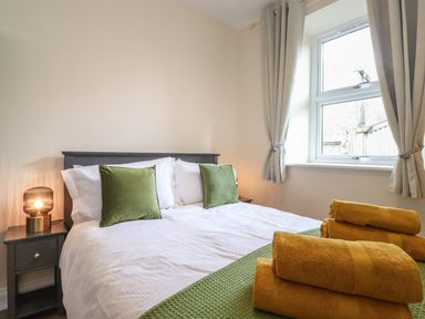 A bedroom with a bed with green cushions and folded yellow towels near a window with curtains at Pond Cottage in Alnmouth