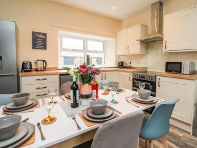 A kitchen with a dining table set for six with bowls plates and glasses a fridge oven microwave and cabinets at Lonnon Cottage in Alnmouth
