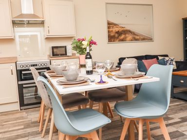 A dining table set for six with bowls plates and glasses in a kitchen next to a sofa with cushions and a framed picture on the wall at Lonnon Cottage in Alnmouth