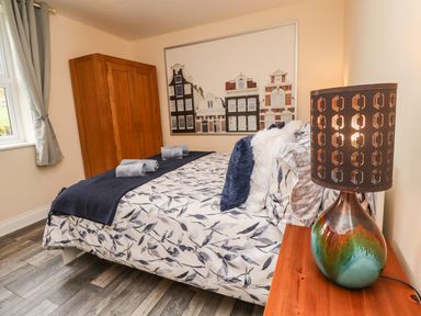 A bedroom with a bed covered with floral bedding and navy linens a wooden wardrobe a window with grey curtains and a colorful lamp on a wooden bedside table at Lonnon Cottage in Alnmouth