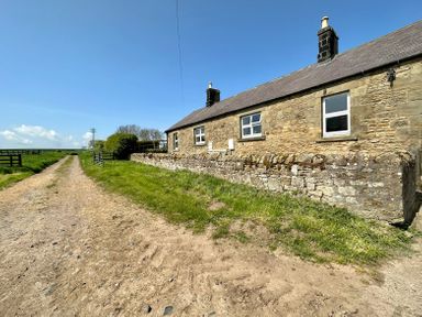 A stone cottage with chimneys next to a dirt path and grassy area at Lonnon Cottage in Alnmouth