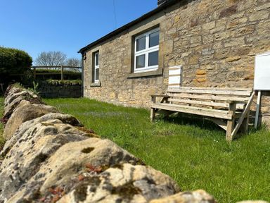 A stone wall garden with grass and a wooden bench next to a stone house with white windows at Lonnon Cottage in Alnmouth