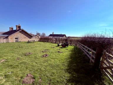 A fenced grassy garden with a picnic table and outdoor chairs near stone houses at Lonnon Cottage in Alnmouth