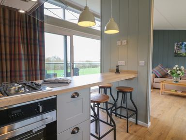 A kitchen area with a stove and oven next to a wooden breakfast bar with three stools and a view of a living room at Willow Lodge in Winthorpe near Newark-On-Trent