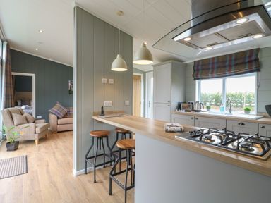 A kitchen with a gas stove and three bar stools at a counter next to a living room with armchairs and a sofa at Willow Lodge in Winthorpe near Newark-On-Trent