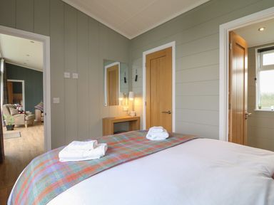 A bedroom with a bed, towels on the bed, a small wooden desk with a mirror and lamps, two wooden doors, and an adjoining living area visible through an open door at Willow Lodge in Winthorpe near Newark-On-Trent