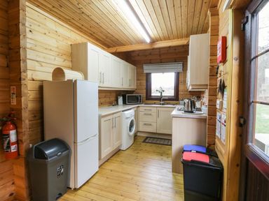 A kitchen with wooden walls and ceiling a refrigerator a washing machine microwave and two trash bins at Moyle Lodge in Dalbeattie