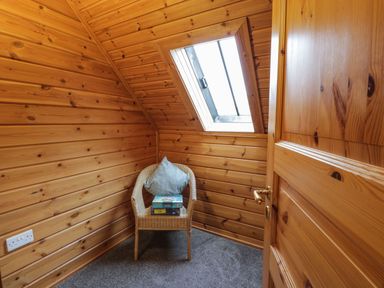 A small wooden room with a wicker chair holding a pillow and board games under a skylight at Moyle Lodge in Dalbeattie